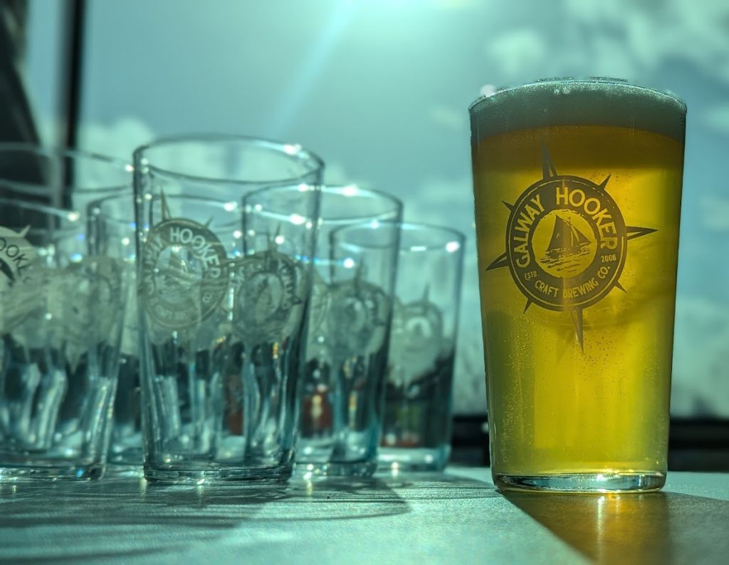 Galway Hooker Craft Beer Glasses in Sunlight – A pint of Galway Hooker craft beer sits on a table, with a row of empty branded glasses behind it, illuminated by bright natural sunlight, creating a crisp and refreshing scene.