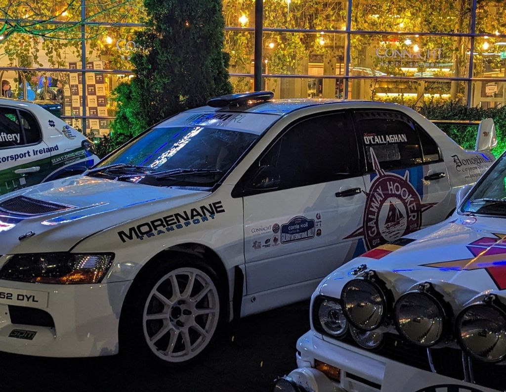 A white rally car with various sponsor logos, including Galway Hooker and Connacht Hospitality Group, parked outdoors at night. The car has "O'Callaghan" on the side window, white rims, and a black roof vent. Other rally cars are partially visible nearby. Warm lights and the illuminated signage of "The Connacht" hotel can be seen in the background.