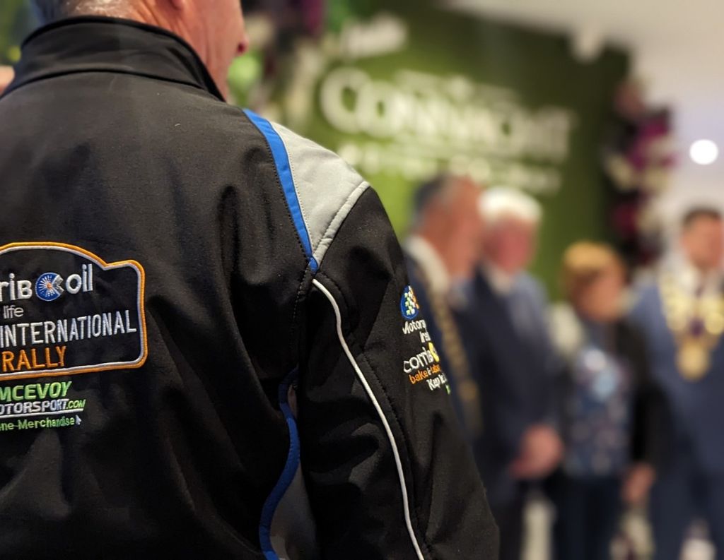 A person wearing a black motorsport jacket with embroidered logos for the Corrib Oil International Rally and the Connacht Hospitality Group. The focus is on the jacket's branding, while a blurred background shows a formal indoor event with people, including local officials, standing in front of a green wall with "The Connacht" branding.