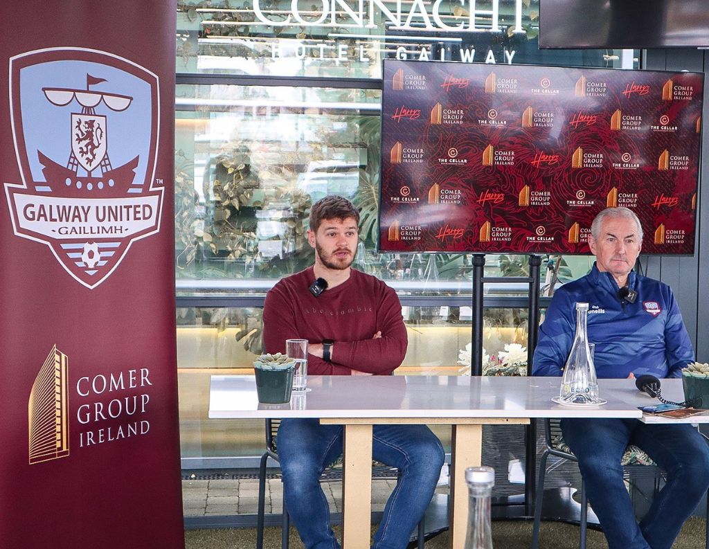 Galway United Conference – Two men sit at a table with microphones in front of a "Galway United" banner and a Comer Group Ireland logo. A screen behind them displays event sponsors, indicating an official team announcement.