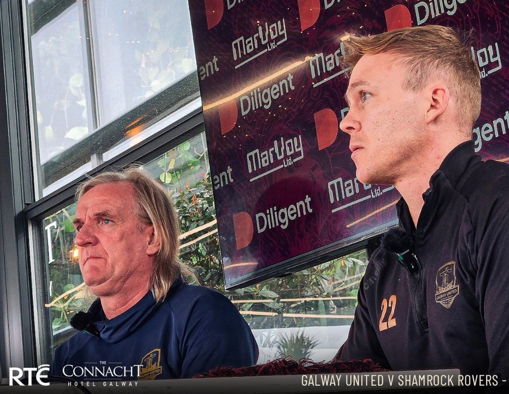 A close-up of two men in Galway United training attire speaking at an event. The background screen shows sponsor logos, suggesting an important discussion related to the team.