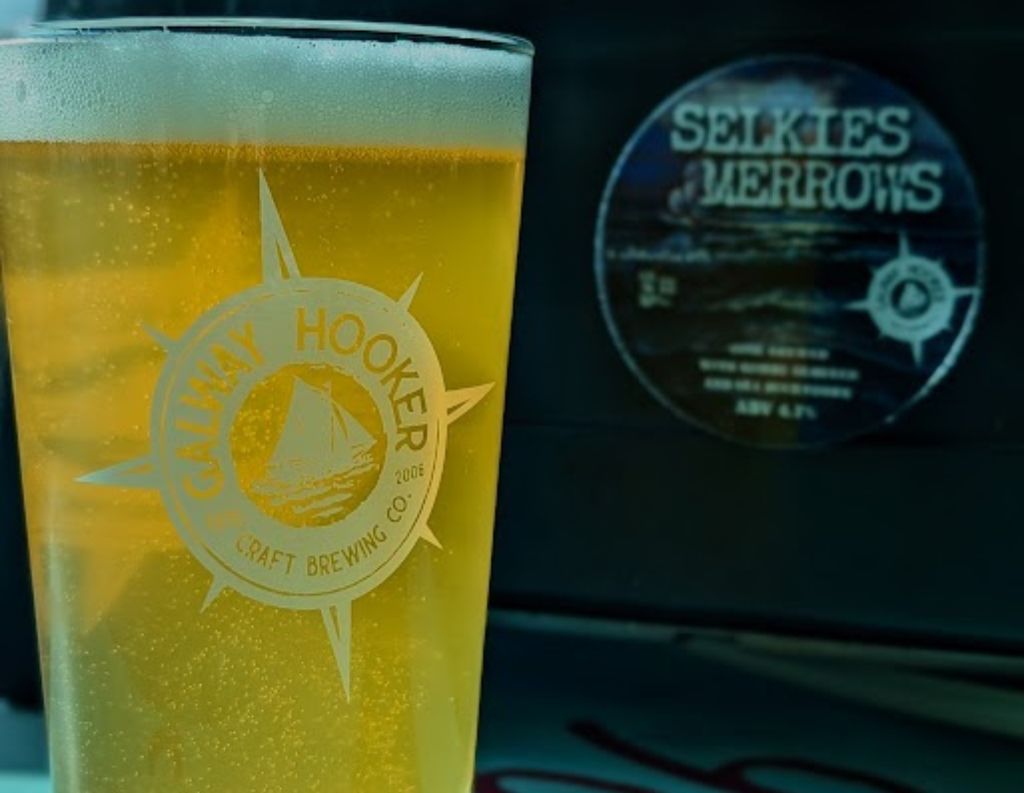 Pint of Galway Hooker Craft Beer with Selkies & Merrows Branding in the Background – A tall glass of golden craft beer with the "Galway Hooker Craft Brewing Co." logo is placed in front of a beer tap labeled "Selkies & Merrows," showcasing a local Irish brew.
