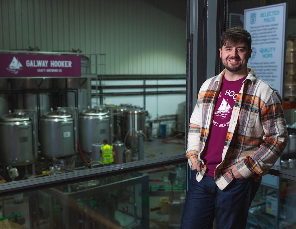 Man Standing in Front of Galway Hooker Brewery Tanks – A young man wearing a Galway Hooker t-shirt and a plaid jacket smiles while leaning on a railing inside a brewery, with large stainless steel fermentation tanks and a "Galway Hooker Craft Brewing Co." sign in the background.