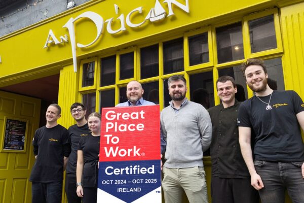 An Púcán Team Holding Great Place to Work Certification – The team of An Púcán, a vibrant pub with a bright yellow exterior, poses cheerfully outside while holding a "Great Place to Work" certification sign for October 2024 - October 2025.