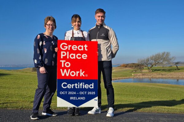 Galway Bay Golf Staff Celebrating Great Place to Work Certification – Three staff members stand on a stunning golf course with a coastal backdrop, dressed in casual and hospitality attire, proudly holding a "Great Place to Work" certification sign for October 2024 - October 2025.