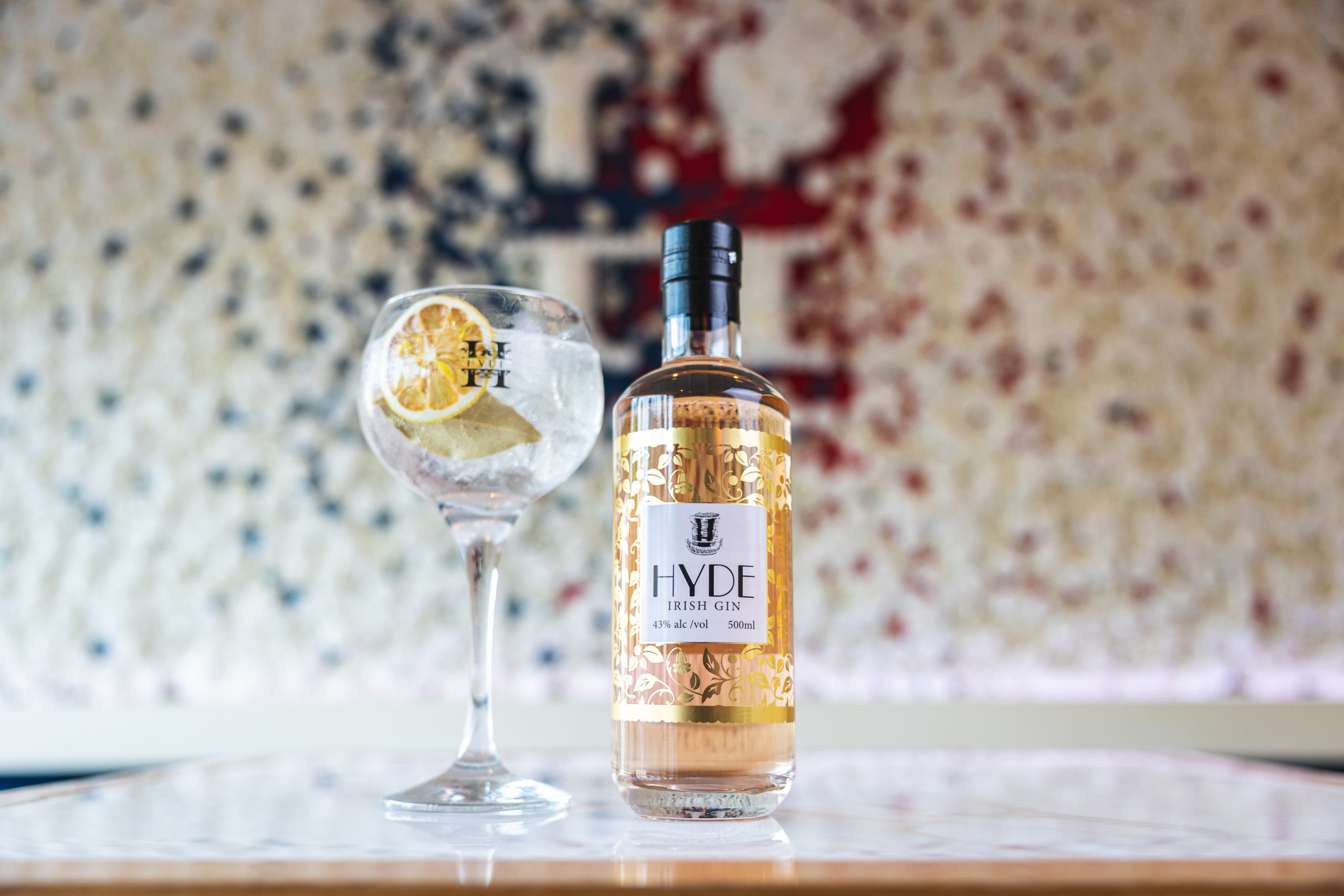 Bottle of HYDE gin next to a filled gin glass with garnish in front of a flower wall with the HYDE logo